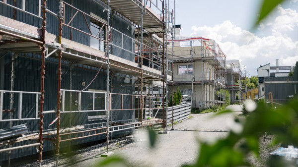 En rad med bolighus under bygging, omgitt av stillaser og byggematerialer, med klar himmel i bakgrunnen. Grønt løvverk er synlig i forgrunnen, noe som gjenspeiler engasjementet for grønn rehabilitering.