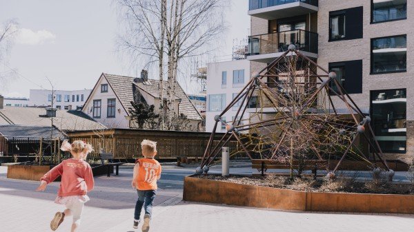 To barn i fritidsklær løper mot en utendørs lekeplassstruktur i et boligområde på en klar dag. Moderne bygårder og trær, som gir inntrykk av et nabolag som gjør seg klar for salg, står stolt i bakgrunnen.